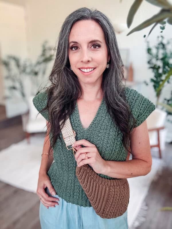 A woman with long dark hair and gray streaks stands indoors, wearing a green knit top, light blue pants, and a Summer House Crochet Cross-Body Bag - Free Pattern in brown. She is smiling and looking at the camera.