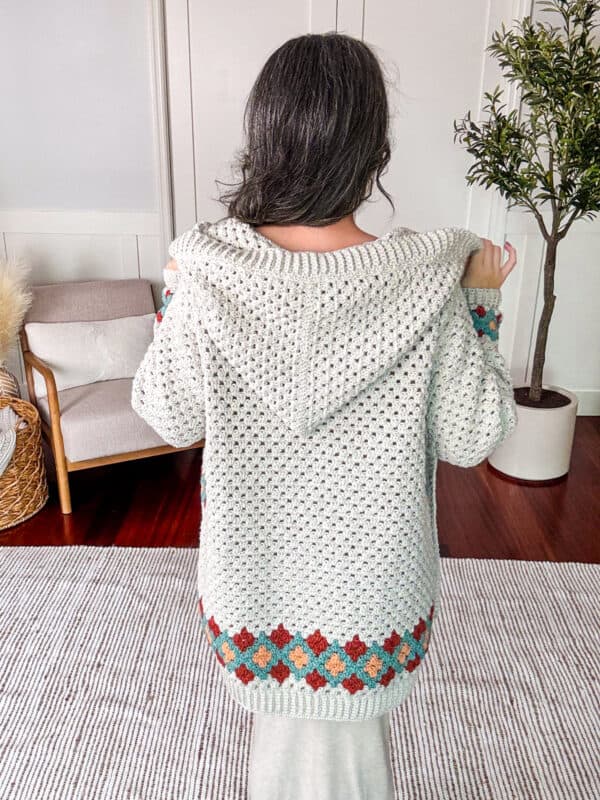 A person with long hair, seen from behind, wearing a white hooded sweater with multicolored accents on the cuffs and hem. They stand in a room with wooden furniture and a plant.