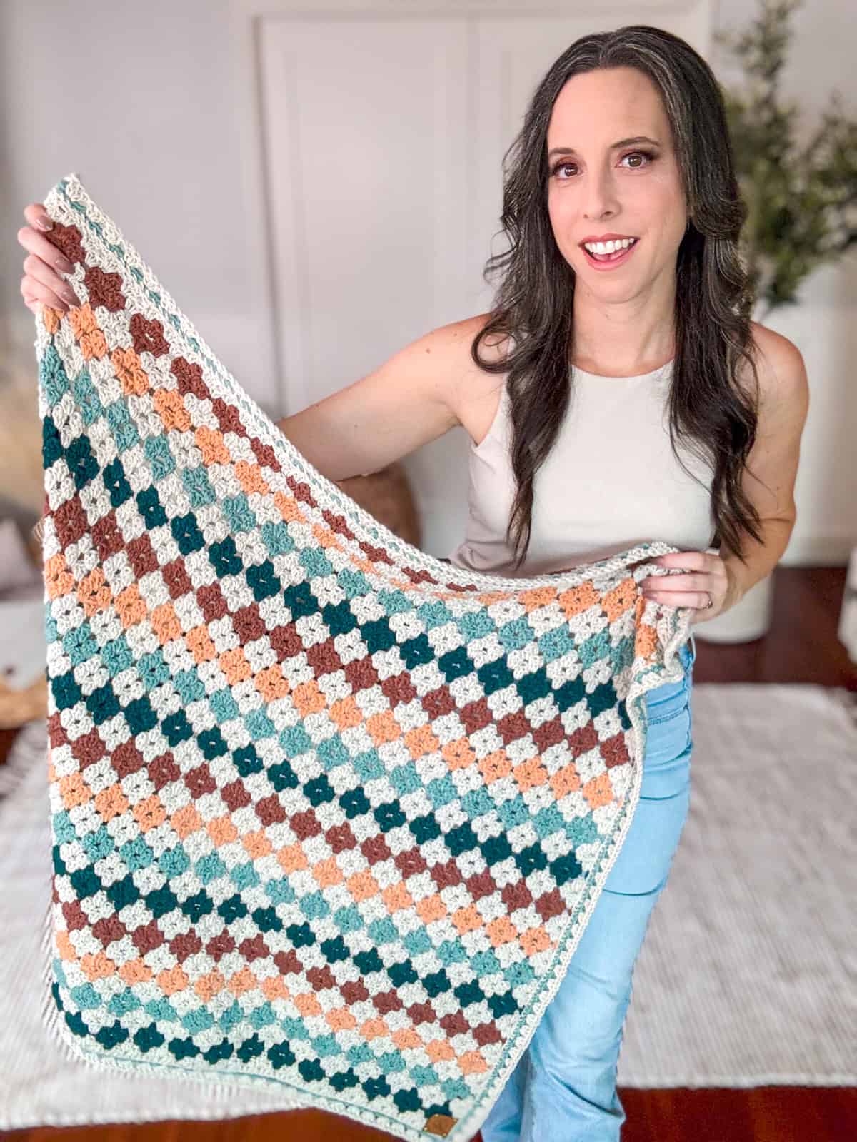 A woman stands indoors holding a crocheted multicolored blanket, featuring a pattern of squares in green, brown, and cream. She wears a light-colored top and blue jeans.