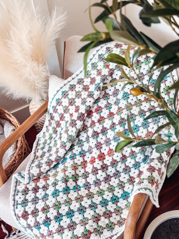 A multicolored crocheted blanket draped over a chair, with a potted plant and ornamental grasses nearby.