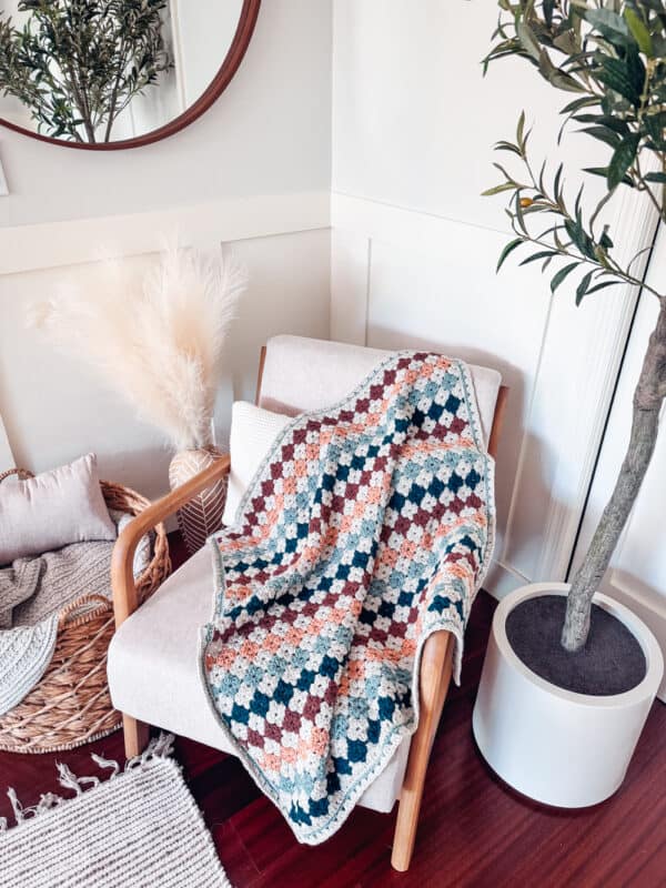 A cozy armchair with a colorful crocheted blanket draped over it sits next to a potted plant and pampas grass, against a white wall with wainscoting.