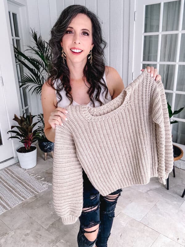 Person smiling and holding up a beige knitted sweater in a room with potted plants and large windows in the background. The cozy Dove Pullover - an easy crochet sweater pattern - complements the serene setting beautifully.