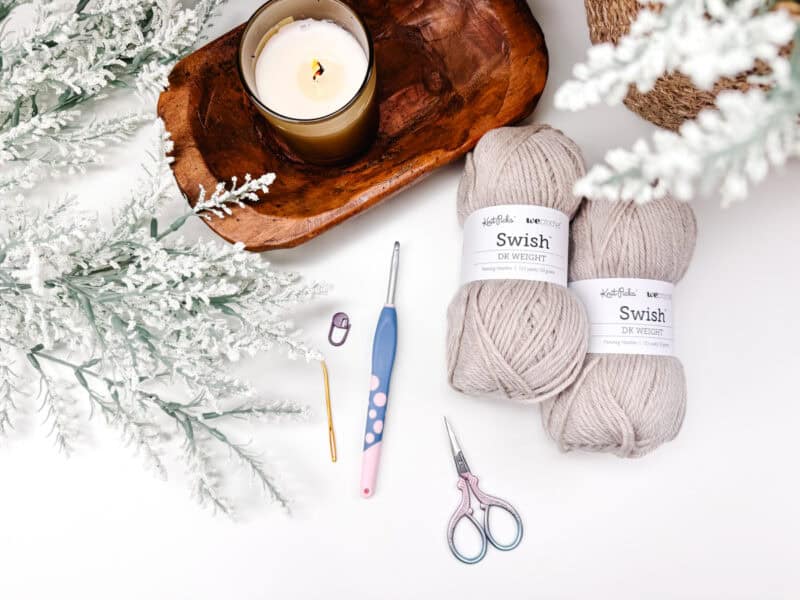 A lit candle, two skeins of beige yarn labeled "Swish," a crochet hook, a tapestry needle, scissors, and a stitch marker are arranged on a white surface with decorative snowy foliage—perfect for working on your Dove Pullover - Easy Crochet Sweater Pattern.