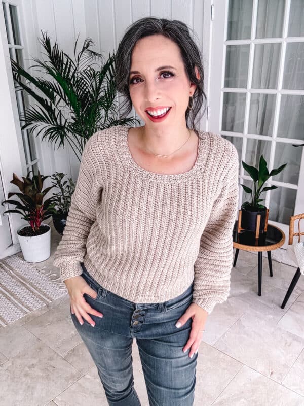 A woman with dark hair smiles at the camera, wearing a Dove Pullover - Easy Crochet Sweater Pattern in beige and blue jeans. She is standing indoors near several potted plants, white walls, and glass-paneled doors.