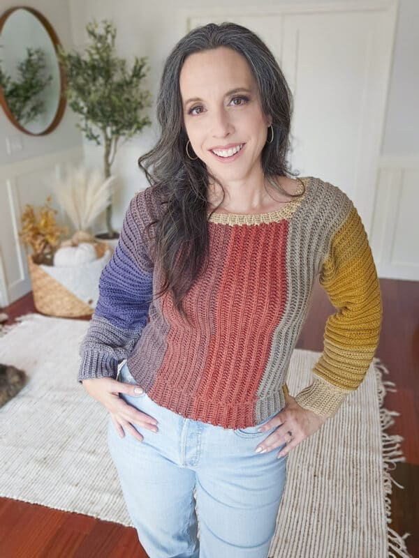 Indoors, a person dons a vibrant Dove Pullover, showing off an easy crochet sweater pattern. The colorful design pairs perfectly with light blue jeans, while a basket and plants add charm to the background.