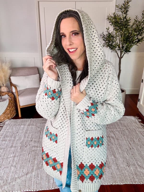 A woman stands indoors wearing a gray crocheted hooded cardigan with colorful patterns. She is holding the hood with one hand and smiling.