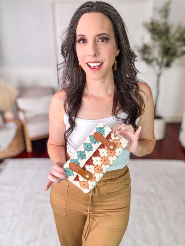 A woman stands indoors holding a crocheted, multicolored handbag. She is wearing a white top and brown pants. The background features a plant and light-colored furniture.