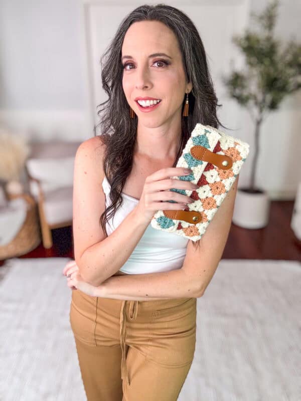 A person with long hair smiles while holding a textured, multicolored clutch bag. They are dressed in a white tank top and tan pants, standing in a room with minimalistic decor and a plant.