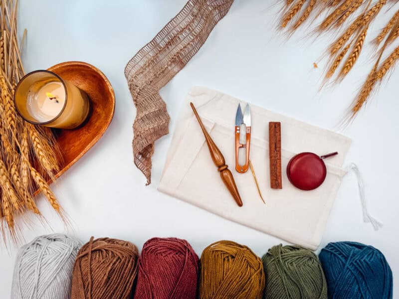 Assorted crochet tools and yarns in various colors are arranged neatly on a white surface, alongside wheat stalks, a candle, and a small wooden tray.