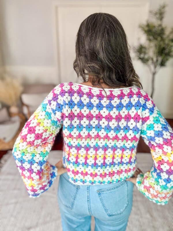 A woman with long brown hair, seen from the back, wearing a colorful crocheted sweater and blue jeans.