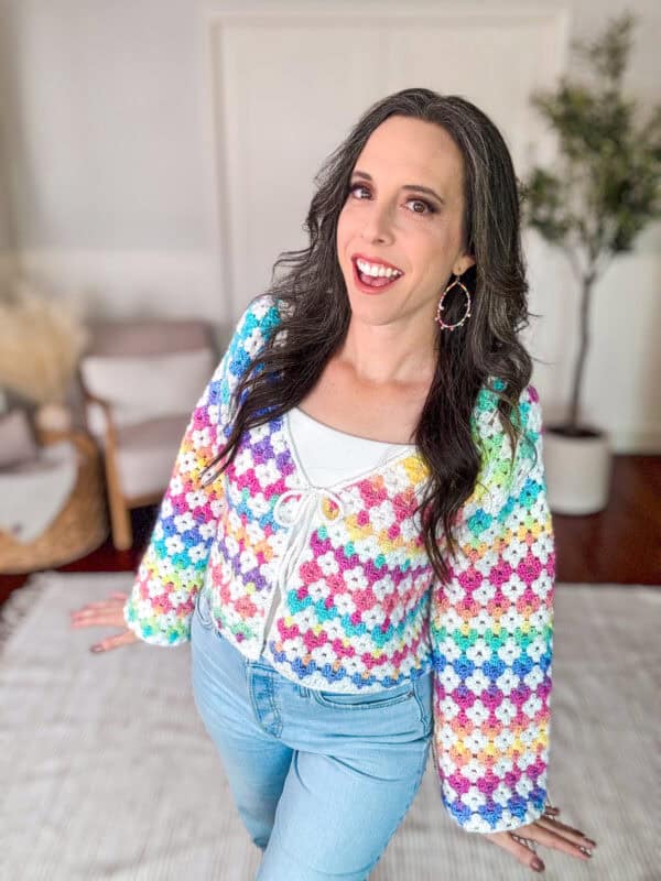 A smiling person wearing a colorful, crocheted cardigan and light blue jeans is standing in a bright room with white walls and a plant.