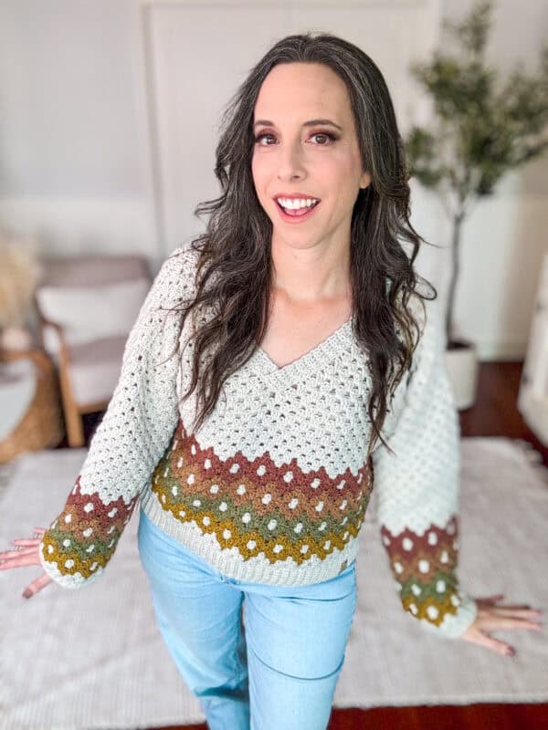 A woman with long dark hair is smiling and wearing a white crocheted sweater with multicolored stripes and light blue jeans. She stands indoors in a room with a plant and furniture in the background.