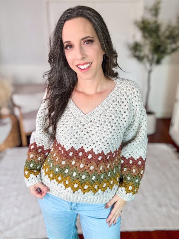 A woman with long dark hair wears a colorful, crocheted sweater with a gradient pattern, standing in a bright room with white and beige decor.