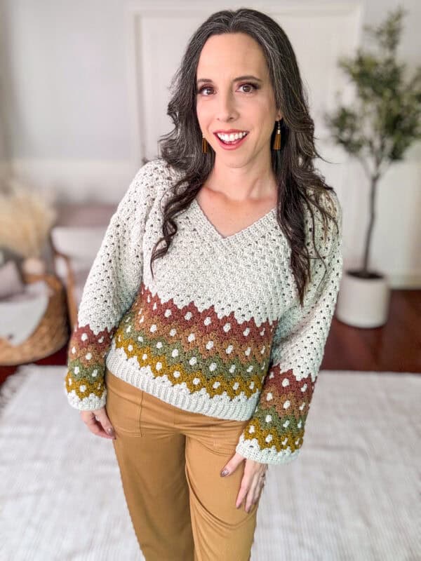 A person with long gray hair stands indoors, smiling and wearing a knitted sweater with a gradient pattern and khaki pants.