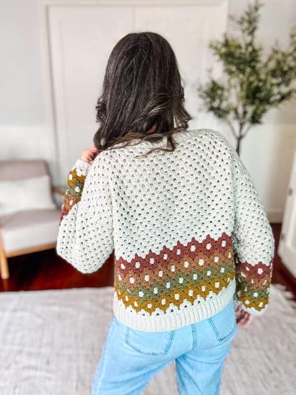 A person with long dark hair is shown from the back wearing a crocheted sweater with multicolored stripes and blue jeans. A plant, a chair, and a rug are visible in the background.