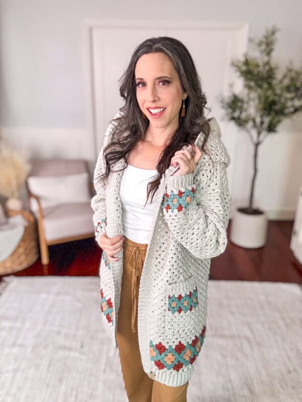 A person standing indoors, wearing a knitted cardigan with colorful patterns, a white top, and brown pants, smiling and holding the cardigan's hood.