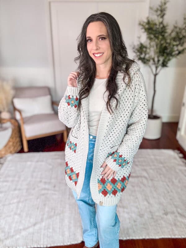 A woman stands indoors wearing a white patterned cardigan and jeans. She smiles at the camera while standing on a light-colored rug with a chair and a plant in the background.