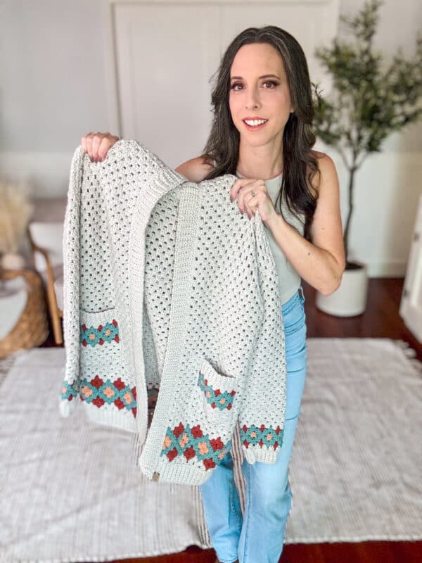 A woman stands indoors, holding up a knitted cardigan with colored patterns on its pockets and lower edges. She is smiling and wearing a light-colored top and blue jeans.