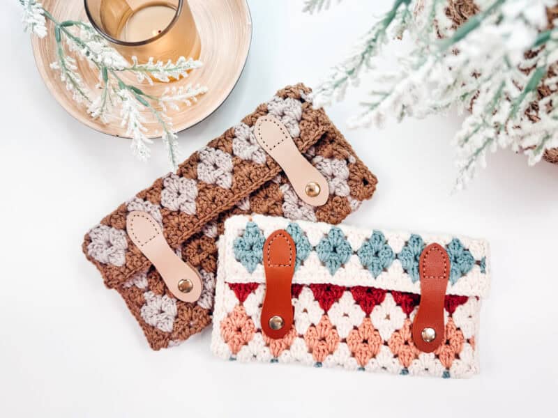 Two crocheted wallets with leather straps lie on a white surface beside a glass of drink and snowy pine branches. One wallet is brown and beige, the other is multicolored.