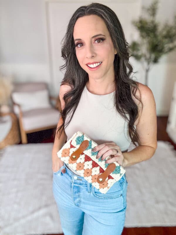 A woman standing in a room holding a crocheted bag, wearing a white sleeveless top and blue jeans, with two chairs and a plant in the background.