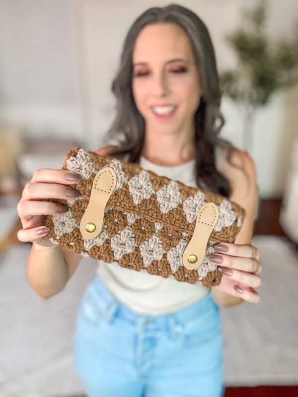 A woman is holding a crocheted clutch purse with a buttoned flap. She has long dark hair and is wearing a white sleeveless top and blue jeans. The background is a blurred indoor setting.