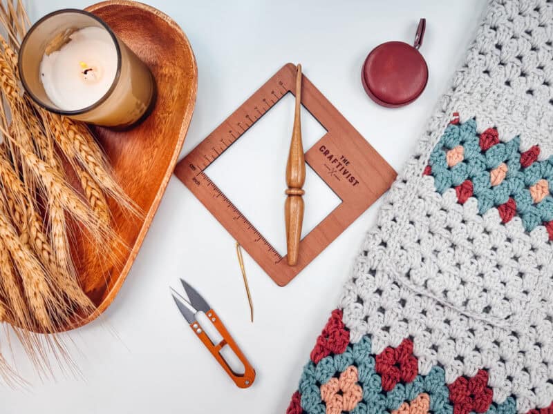 A crocheted blanket with a colorful pattern, a wooden crochet hook, a square measuring tool, a lit candle, wheat stems in a wooden tray, a round tape measure, and orange scissors on a white surface.