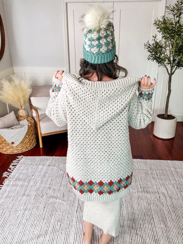 A person facing away and standing in a room, wearing a knitted hat and a matching crocheted cardigan with colorful patterns near the hem. The room has a chair, a basket, and a plant.