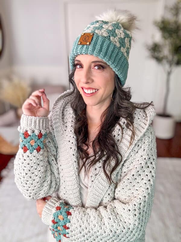 A woman wearing a knitted hat with a pom-pom and a patterned sweater smiles at the camera indoors.