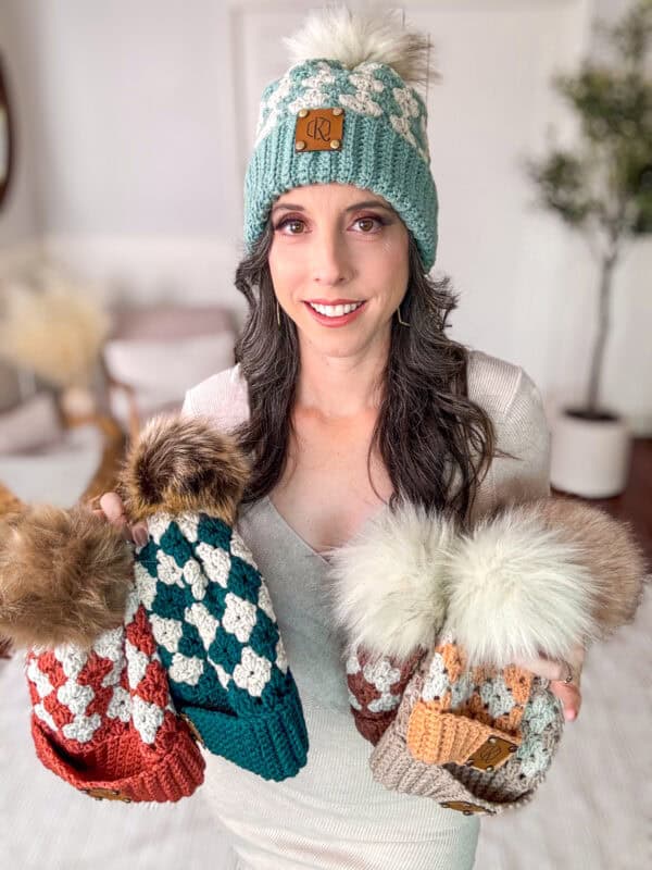 A woman in a white top holds three Diamond Granny Stitch Crochet Hats with fur pom-poms. She wears a similar hat with a pom-pom and a brown tag. The background features cozy home decor.