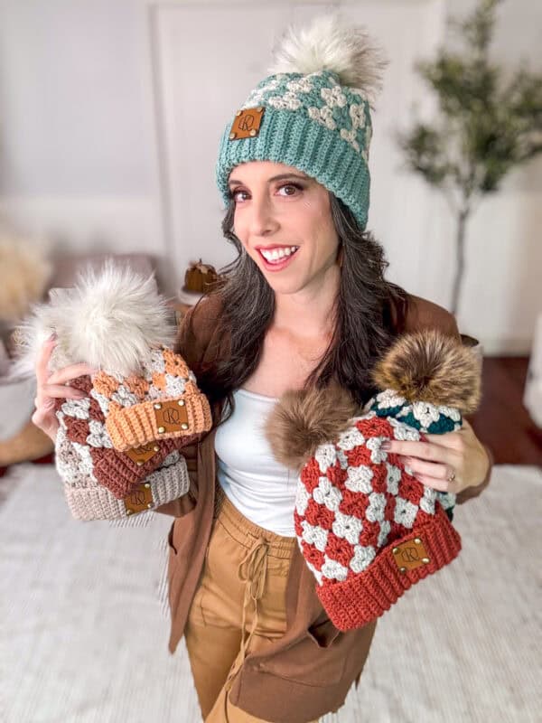 A woman wearing a teal and white knit hat holds four other knit hats with pom-poms, each in a different color pattern.