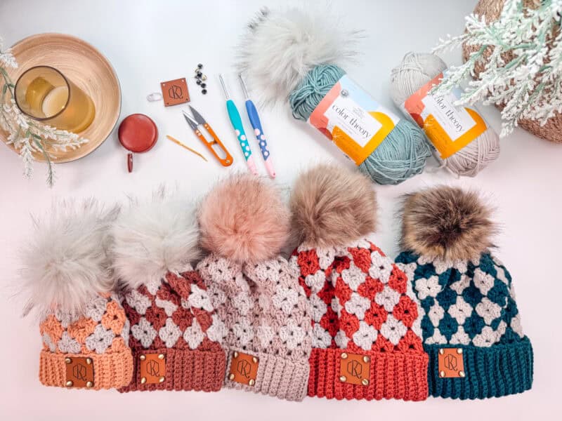 A variety of colorful knitted hats with large pom-poms are displayed alongside crochet hooks, small scissors, a cup of tea, and two skeins of yarn on a white surface.