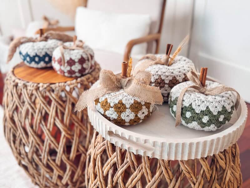 Handmade crocheted pumpkins with burlap ribbons are displayed on woven tables in a bright room.