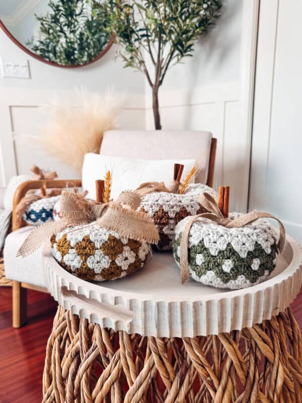 A decorative tray on a wicker table features three crocheted pumpkins with cinnamon stick stems and burlap bows. In the background, a white armchair and a potted plant are visible.