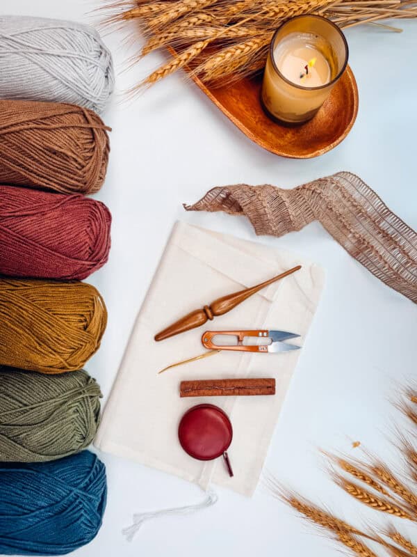 A collection of yarn balls in different colors, a lit candle, wheat stalks, and various knitting tools are arranged on a white surface.