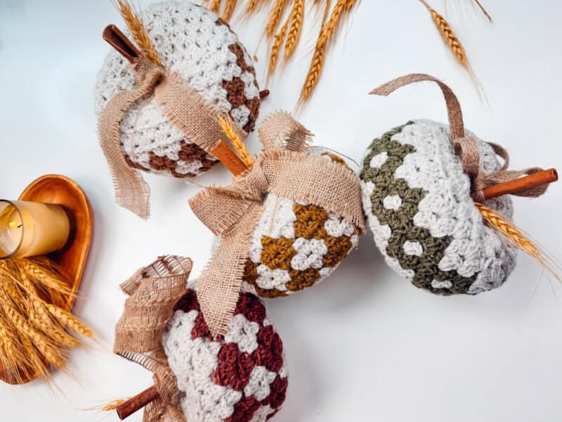 Four crocheted pumpkins in various patterns with cinnamon stick stems and burlap ribbons, placed on a white surface with wheat stalks and a lit candle on a wooden tray in the background.