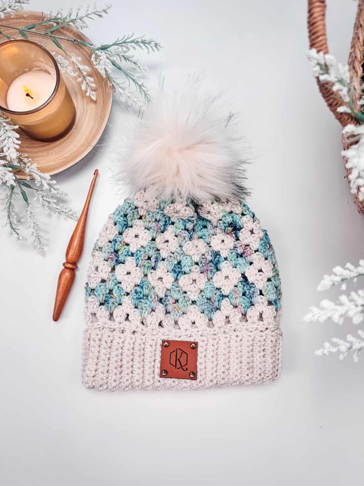 A Diamond Granny Stitch Crochet Hat with a multicolored pattern and a fluffy pom-pom, displayed alongside a wooden crochet hook, a lit candle, and decorative foliage.