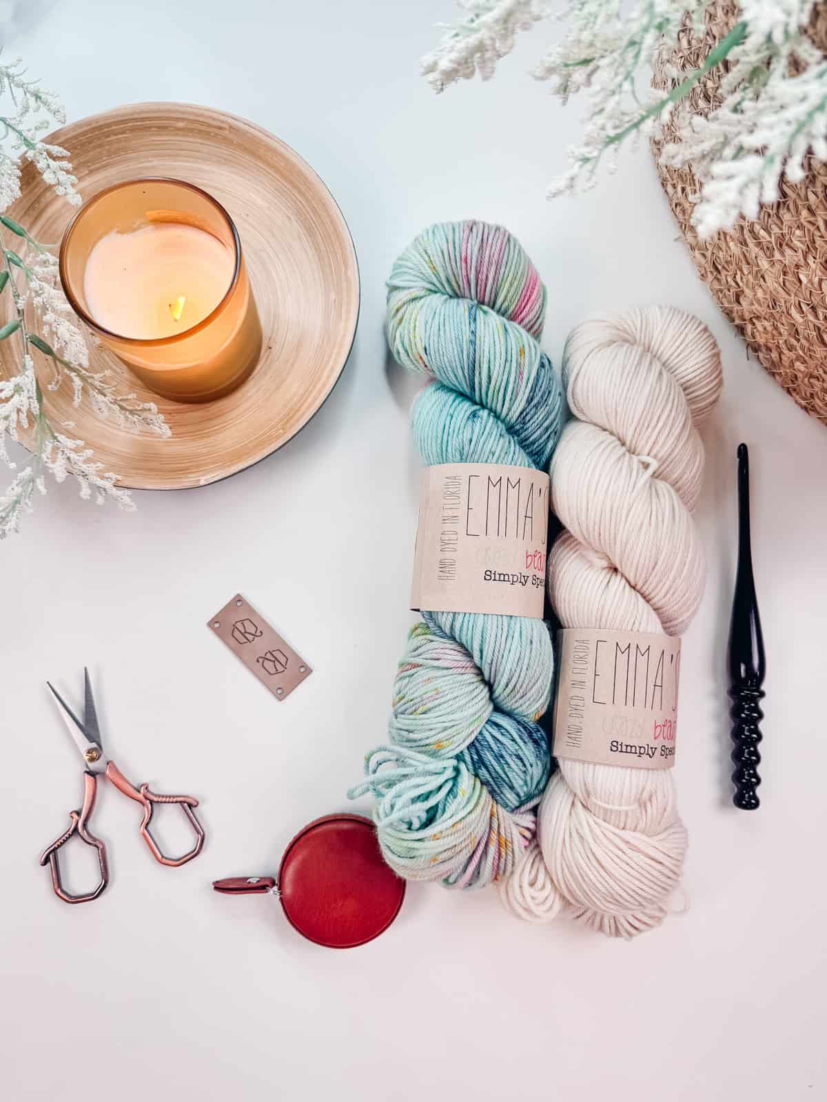 A tabletop with two skeins of yarn, a lit candle, scissors, crochet hook, a leather tape measure, and some small metal labels all set for crafting a beautiful Diamond Granny Stitch Crochet Hat. A potted plant is partially visible in the corner.