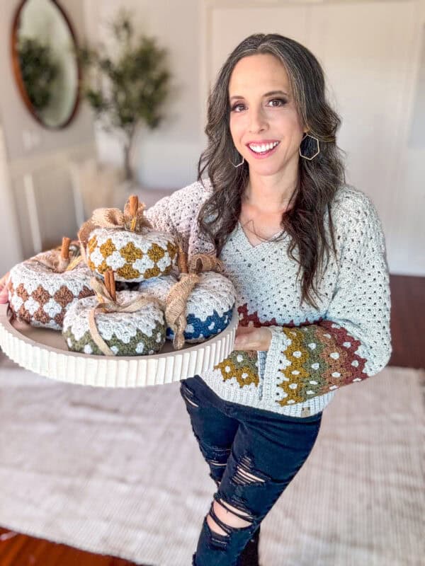 A woman with long hair, wearing a Diamond Granny Stitch crochet-patterned sweater, holds a tray of decorative knitted pumpkins. She stands indoors in a room with a mirror and a plant in the background.