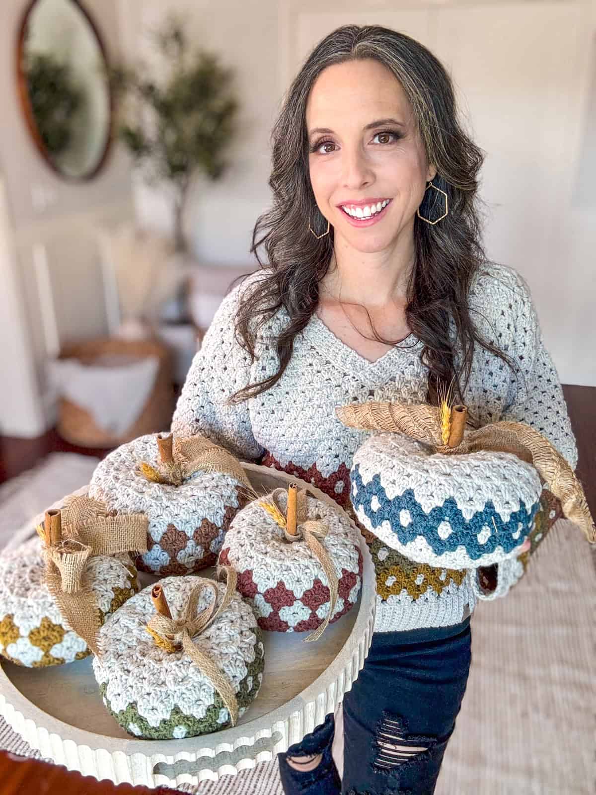 A woman holding a tray with six handmade Diamond Granny Stitch Crochet Pumpkins of different colors stands in a room decorated with plants and a mirror. She is smiling and wearing a light-colored sweater.