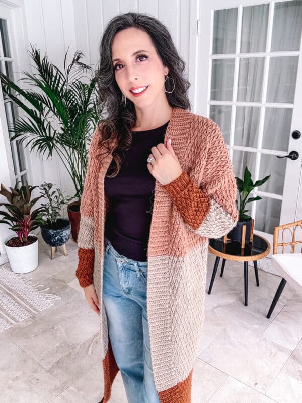 A person with long hair wearing a brown and beige knit cardigan and jeans stands indoors, near plants and a glass-paneled door.