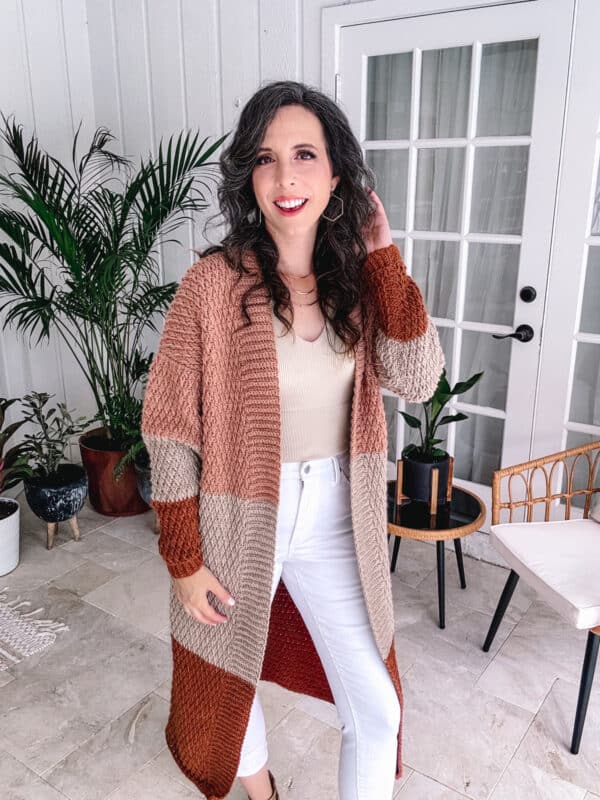 Person standing indoors, wearing a long color-block cardigan with rust, beige, and cream tones, a cream top, and white pants. There are potted plants and a chair in the background.