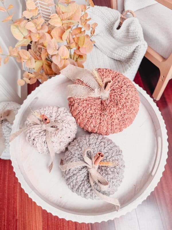 A wooden tray holds three knitted pumpkins in orange, grey, and beige tones. The background includes a knitted blanket, dried leaves, and wooden furniture.