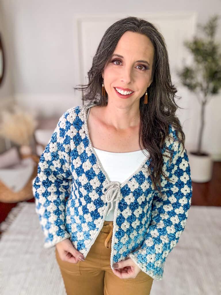 A person with long, dark hair is smiling and wearing a blue and white patterned cardigan over a white top and brown pants, standing in a room with a plant and a mirror in the background. The cozy setting highlights her Rainbow Hues Granny Stitch Crochet Cardigan beautifully.