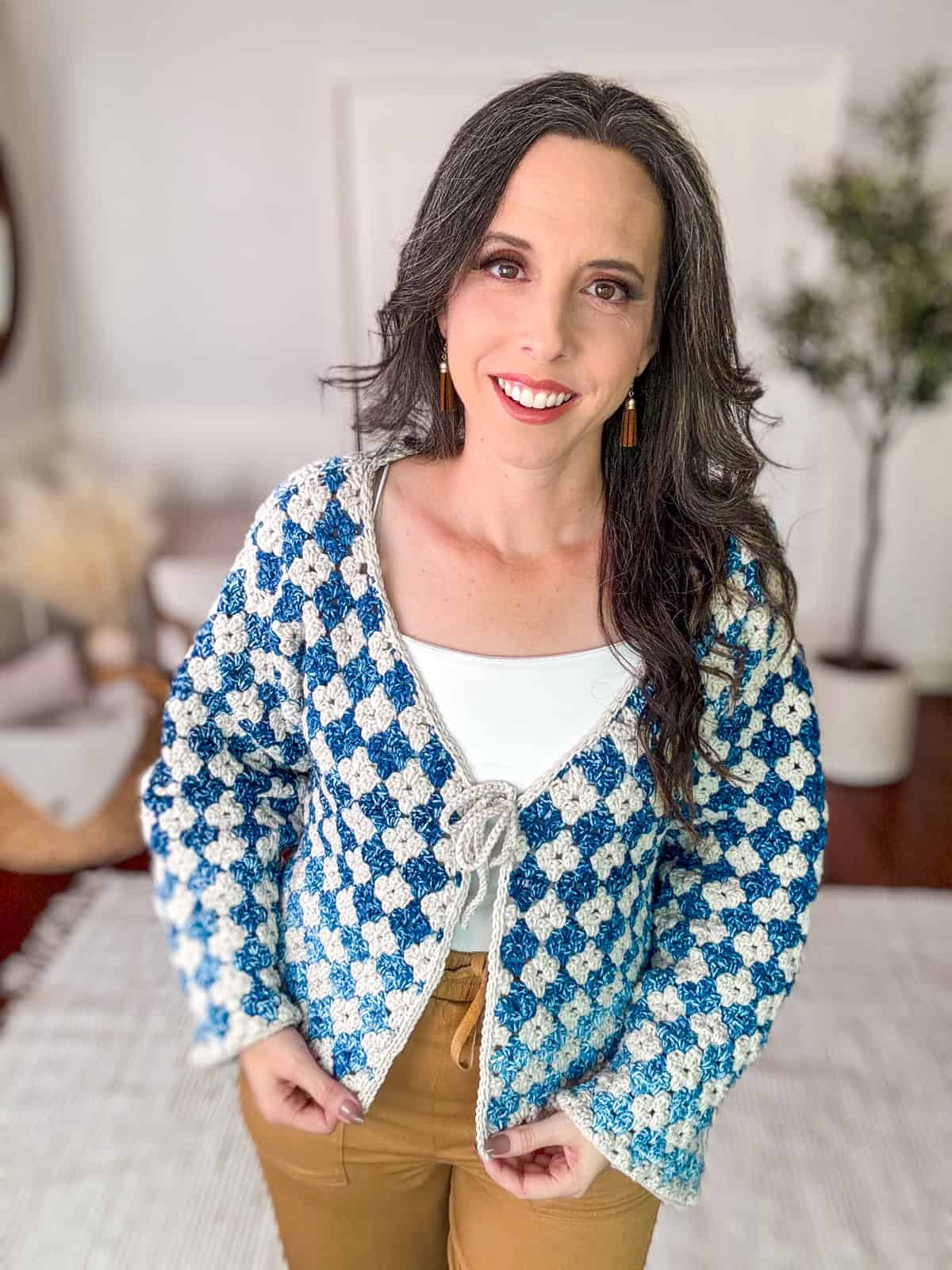 A person with long, dark hair is smiling and wearing a blue and white patterned cardigan over a white top and brown pants, standing in a room with a plant and a mirror in the background. The cozy setting highlights her Rainbow Hues Granny Stitch Crochet Cardigan beautifully.