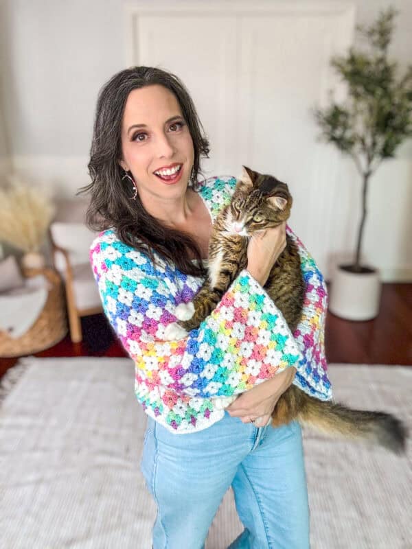 A person with long dark hair, wearing a colorful crochet sweater, holds a tabby cat indoors. There is a plant and a wicker basket in the background.