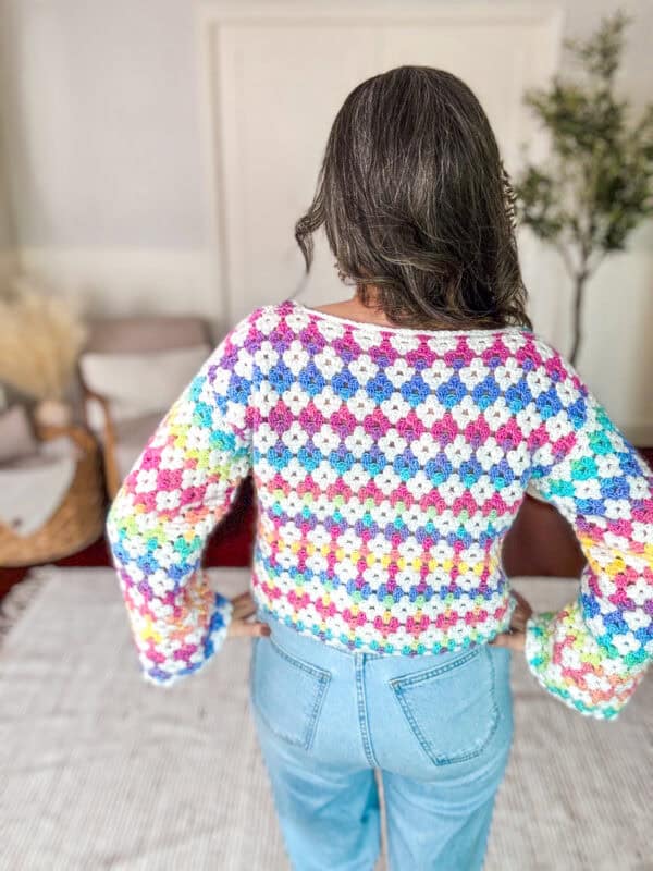 A person with long hair faces away, wearing a multicolored, crocheted sweater with diamond patterns and high-waisted light blue jeans.