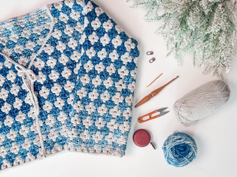 A blue and white crochet blanket is displayed with two crochet hooks, stitch markers, scissors, measuring tape, and two yarn balls on a white surface next to greenery.