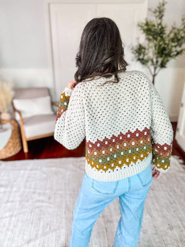 A person with long hair in a patterned sweater and blue jeans stands in a room with a chair, woven basket, plant, and rug. They are facing away from the camera.