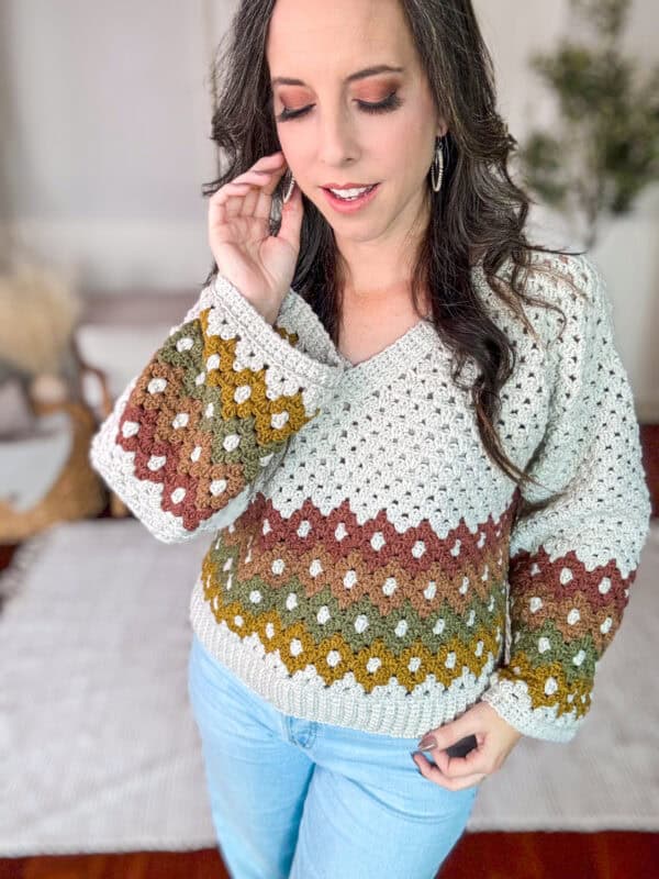 A woman wearing a multicolored crochet sweater and blue jeans touches her hair while standing indoors.