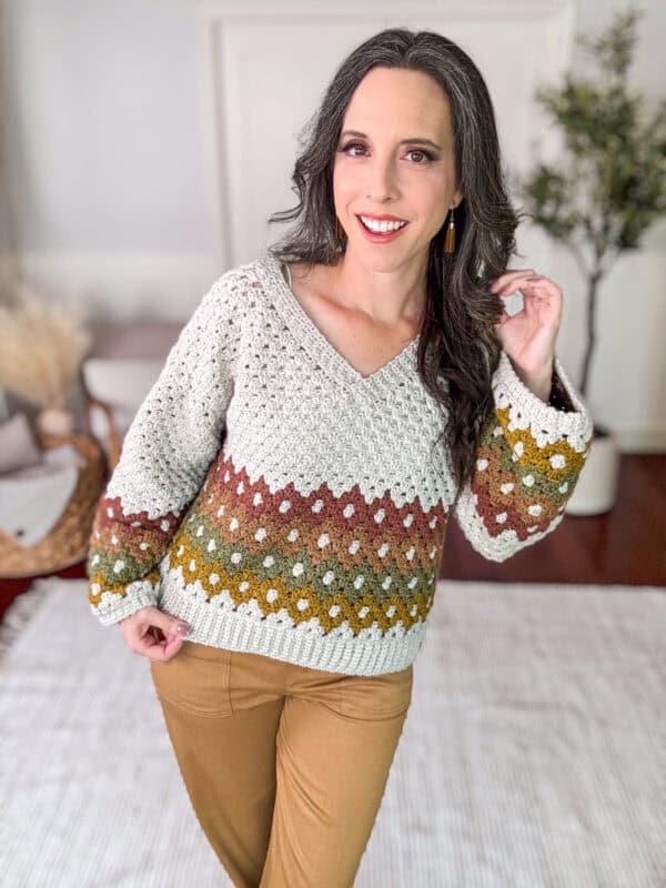 A woman stands indoors wearing a knit sweater with a V-neck and earthy tones of green, brown, and white, paired with camel-colored pants. She has long dark hair and smiles at the camera.